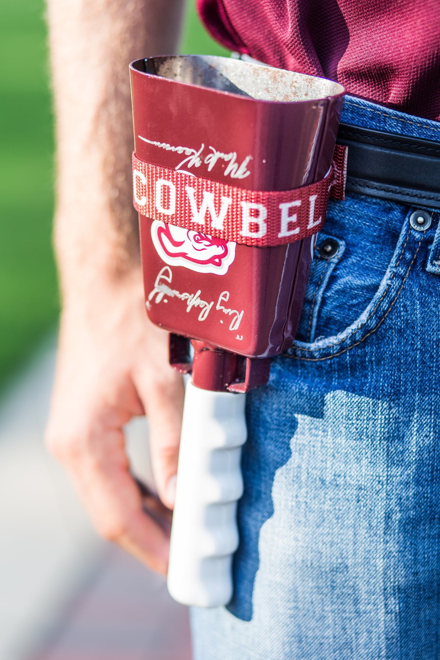 Maroon Cowbelt holstering a cowbell with white handle held by a person wearing blue jeans.